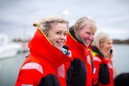 Tres estudiantes de Bexhill College llevan chalecos salvavidas rojos brillantes y están juntos frente a un cuerpo de agua bajo un cielo nublado.