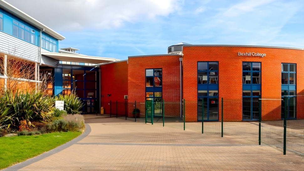El moderno edificio de ladrillo de Bexhill College con grandes ventanas y un camino hacia la entrada rodeado de áreas verdes.