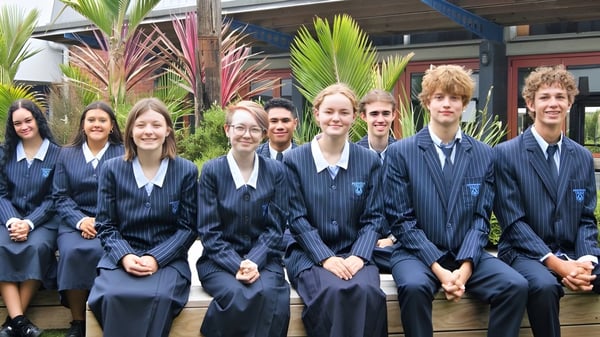 Un grupo de alumnas y alumnos en uniforme escolar está frente al edificio del Birkenhead College con plantas tropicales de fondo.