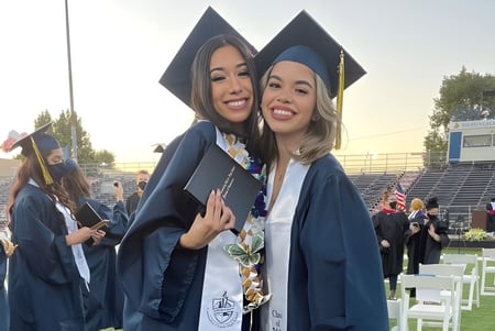 Dos jóvenes mujeres en togas de graduación están juntas frente a un estadio en el terreno de la Birmingham Community Charter High School.