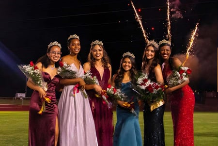 Estudiantes de la Bishop Alemany High School están en vestidos festivos con ramos de flores en el campo de deportes mientras se ve un espectáculo de fuegos artificiales en el fondo.