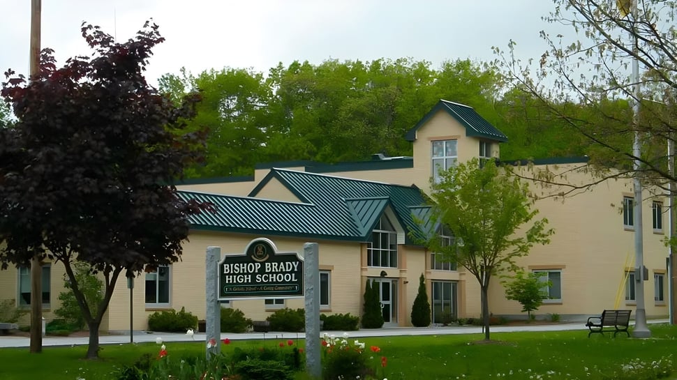El edificio de ladrillo de dos pisos con techo de metal verde pertenece a la Bishop Brady High School y está rodeado de árboles y césped.