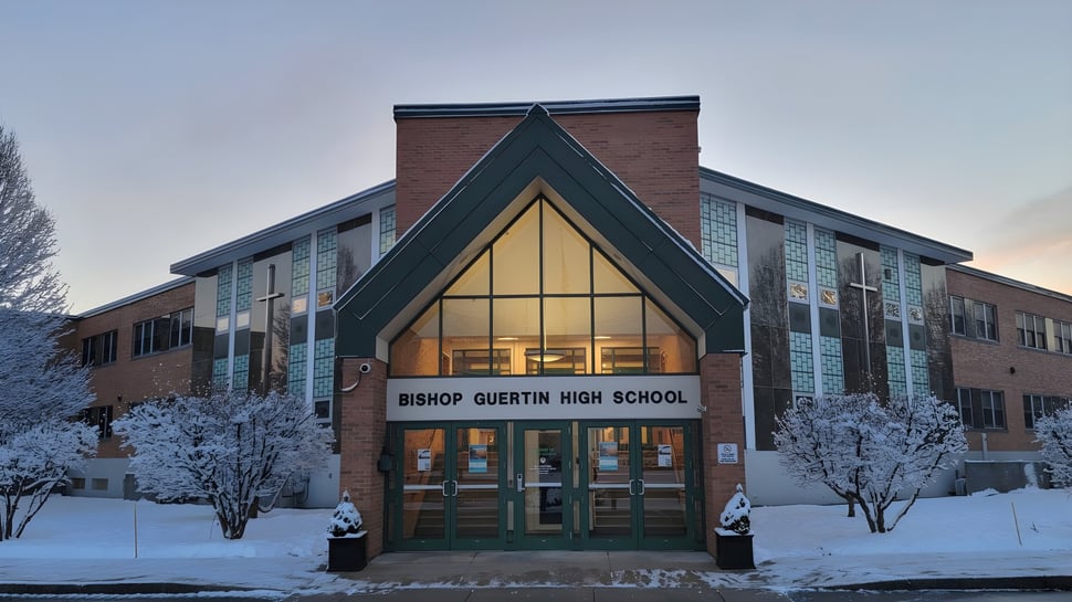 El edificio de ladrillo de la Bishop Guertin High School con techo a dos aguas está rodeado de árboles cubiertos de nieve.