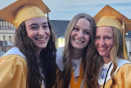 Tres estudiantes en togas de graduación están frente a un edificio en el campus de la Bishop Guilfoyle Catholic High School.