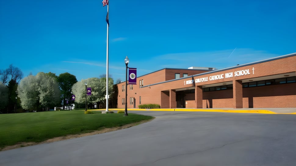 El edificio de ladrillo de la Bishop Guilfoyle Catholic High School con un asta de bandera frente a un campo y árboles bajo un cielo azul.