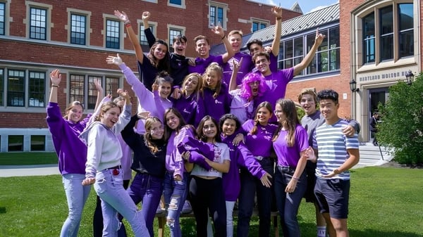 Un grupo de estudiantes en ropa morada posan frente a un edificio de ladrillo en el campus de Bishop’s College School.