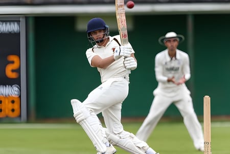 Un jugador de cricket con camiseta blanca está en el bate mientras otro jugador observa en el campo del Bishop’s Stortford College.