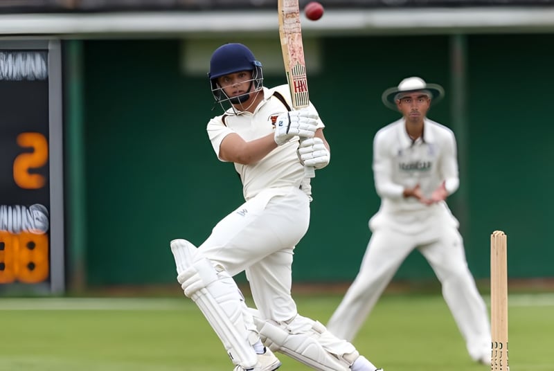 Un jugador de cricket con camiseta blanca está en el bate mientras otro jugador observa en el campo del Bishop’s Stortford College.