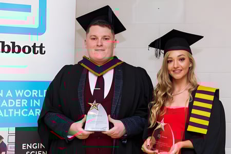 Dos graduados en togas están frente a un logo de Abbott en el campus de la Blackwater Community School.