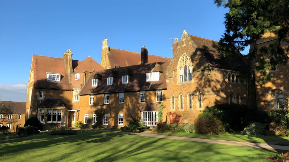 El histórico edificio de ladrillo de la Bloxham School está rodeado de césped bien cuidado y áreas verdes bajo un cielo despejado.