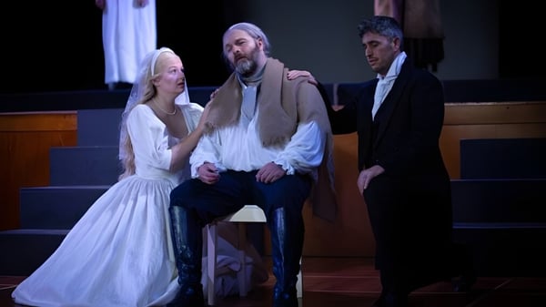 Una mujer con vestido blanco y dos hombres con trajes están juntos en el escenario en Blue Mountain College.
