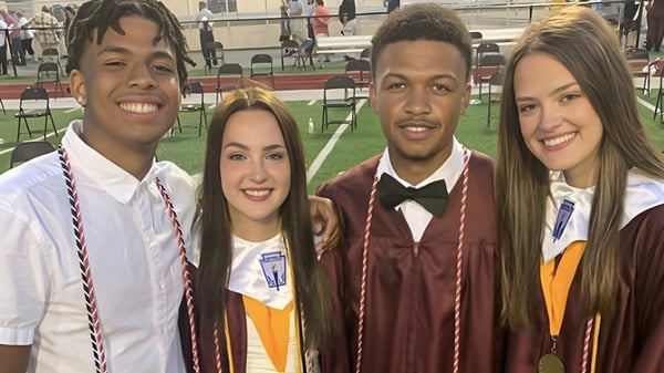 Cuatro alumnas y alumnos de Bluefield High School están juntos sonriendo en una ceremonia de graduación frente al escenario.