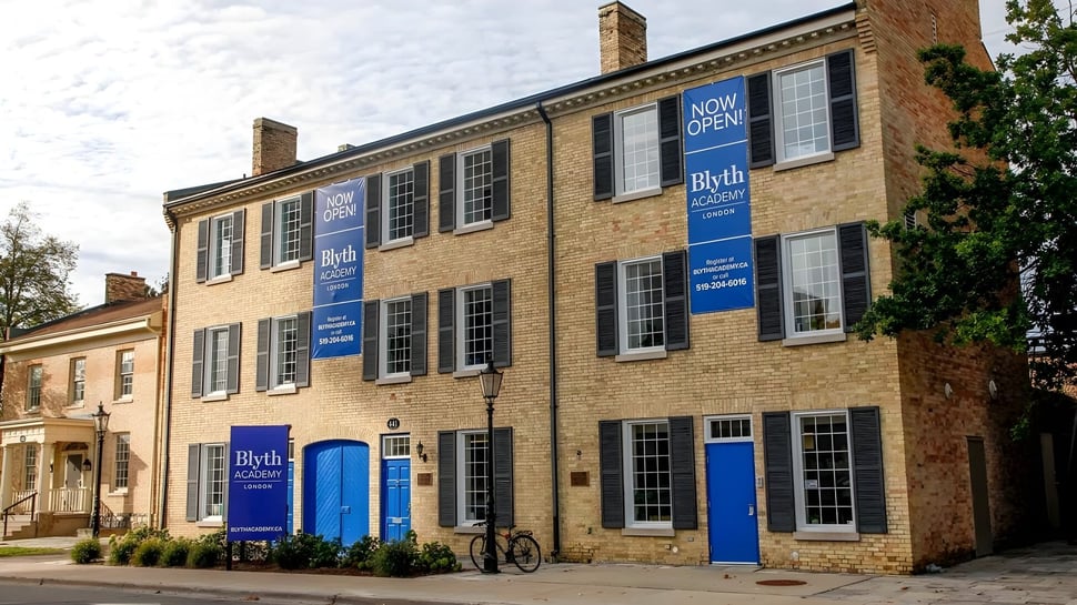 El edificio de ladrillo de varios pisos de la Blyth Academy con ventanas y puertas azules está abierto y rodeado de árboles.