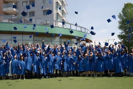 Un gran grupo de graduadas y graduados de la Bodwell High School se reúne afuera en togas azules frente a un edificio con árboles.
