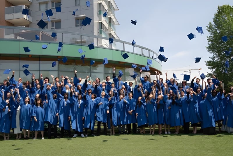 Un gran grupo de graduadas y graduados de la Bodwell High School se reúne afuera en togas azules frente a un edificio con árboles.