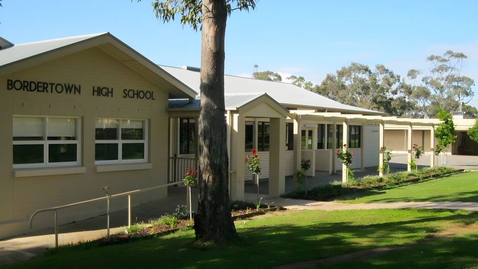 El edificio escolar de una planta de Bordertown High School se encuentra detrás de una palma en un área cubierta de hierba.