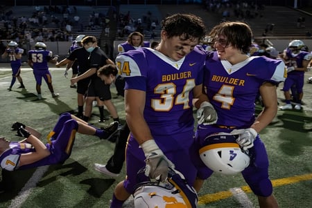 Dos jugadores de fútbol americano están en el campo de Boulder Valley Public Schools con jugadores y espectadores al fondo.