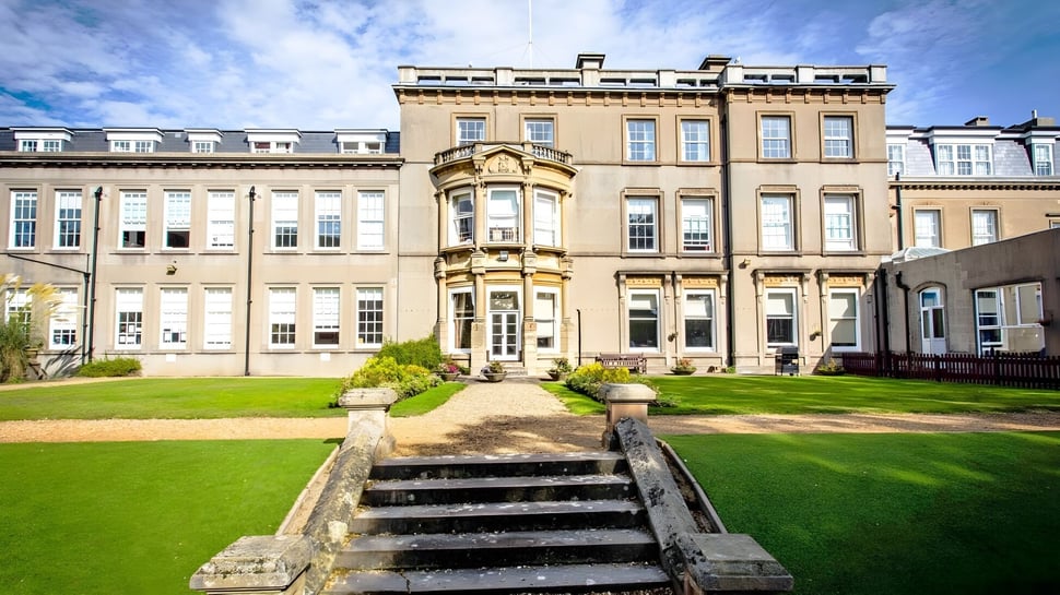 El edificio principal de Bournemouth Collegiate School con césped bien cuidado y escaleras de piedra bajo un cielo azul.