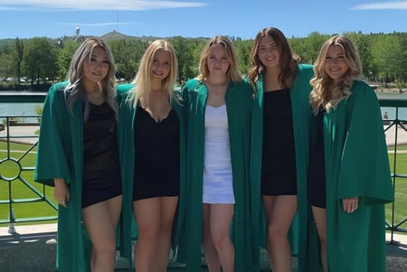 Un grupo de cinco estudiantes en togas de graduación verdes está en el campus de la Bowness High School frente a un lago y montañas.