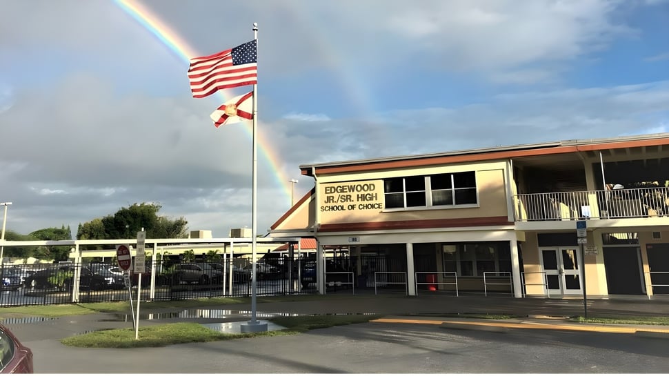 Delante del edificio escolar de Brevard County Public Schools ondean la bandera americana y una bandera estatal bajo un arcoíris en el cielo nublado.