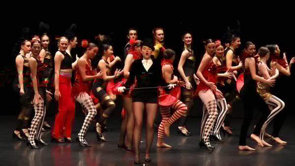 Un grupo de estudiantes del Brighton College presenta una colorida actuación en el escenario con trajes vibrantes en un escenario oscuro.