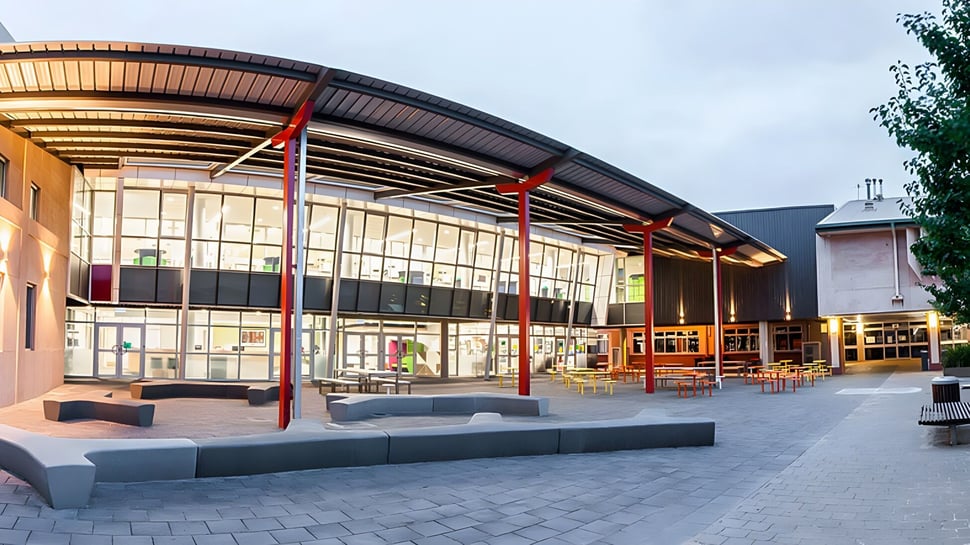 El moderno edificio de la Brighton Secondary School con fachada de vidrio y techo curvado se encuentra frente a un patio pavimentado con bancos y árboles.