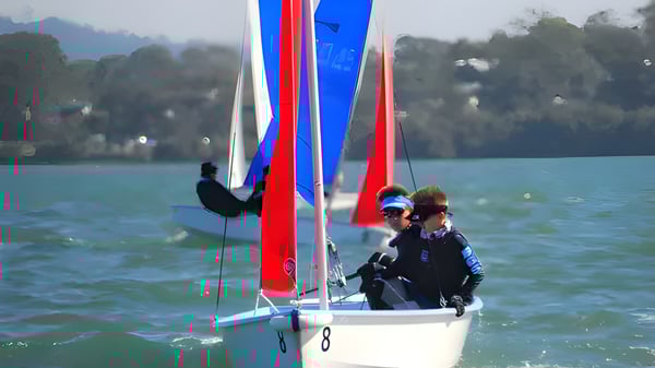 Un velero con velas de colores en un cuerpo de agua frente a montañas y árboles en el terreno de la Brisbane Grammar School.