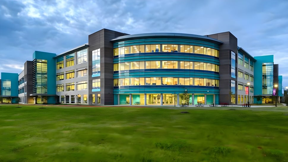 El moderno edificio de varios pisos de la British International School Houston con una fachada de vidrio curvada se encuentra en un prado verde bajo un cielo nublado.