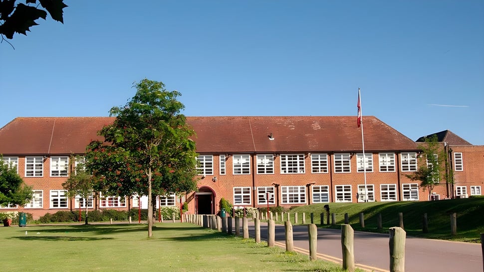 El edificio de ladrillo de varios pisos con techo rojo del Brockenhurst College se encuentra en un área verde con árboles y cielo azul.