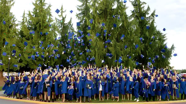 Un grupo de graduados en togas azules se encuentra frente a altos árboles perennes en el terreno de la Brookswood Secondary School.