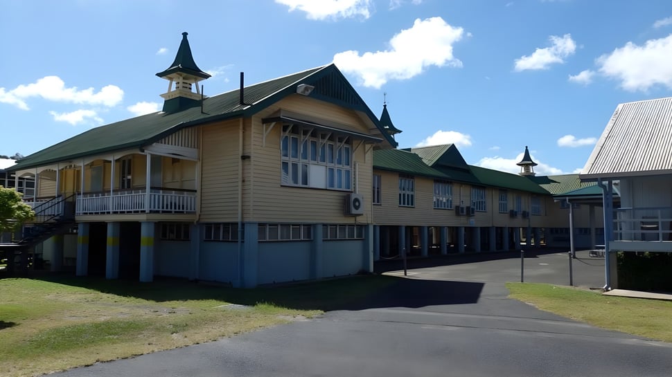 El edificio de madera de varios pisos de la Bundaberg State High School está rodeado de un área de césped bajo un cielo azul con nubes blancas.