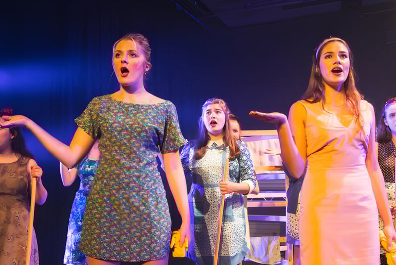 Un grupo de alumnas de Burgess Hill Girls está de pie de manera dramática en el escenario con vestidos coloridos.
