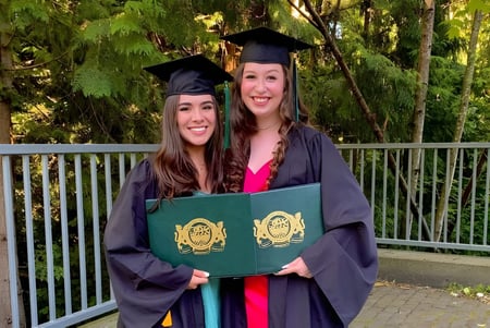 Dos alumnas de la Burnaby Mountain School llevan su vestimenta de graduación y sostienen un diploma en el bosque.
