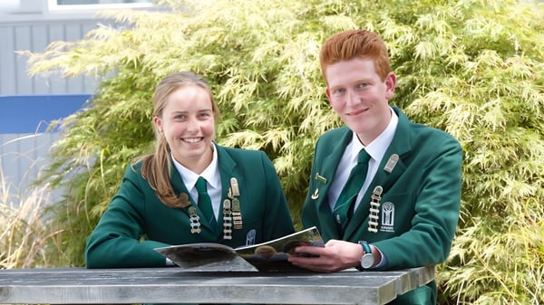 Dos estudiantes en uniformes escolares verdes están juntos en el terreno de Burnside High School frente a un follaje verde.