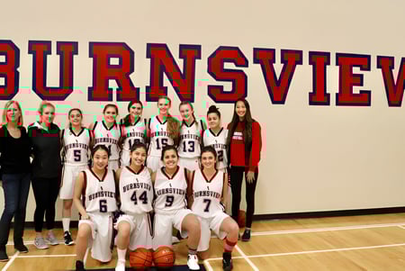 Un grupo de jugadoras de baloncesto posan frente al letrero de la Burnsview Secondary School en el gimnasio.