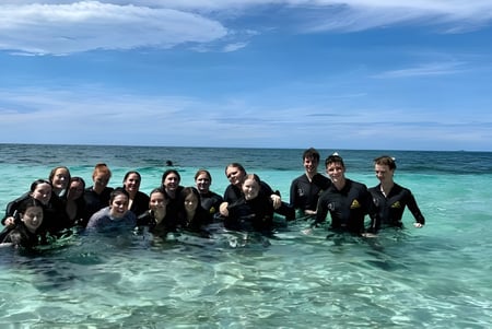 Un grupo de estudiantes del Burpengary State Secondary College está en trajes de neopreno en las aguas poco profundas y turquesas de una playa tropical.
