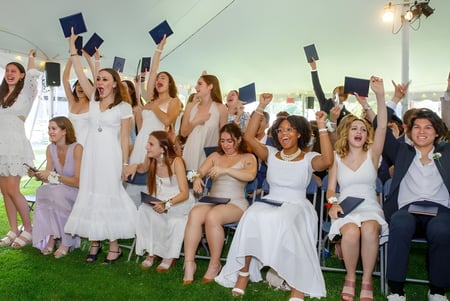Un grupo de alumnas de la Cambridge School of Weston celebra afuera con vestidos blancos y birretes su fiesta de graduación.