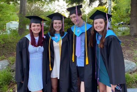 Un grupo de cuatro jóvenes mujeres vestidas de graduación está en el campus de la Camden Hills Regional High School en un entorno verde.