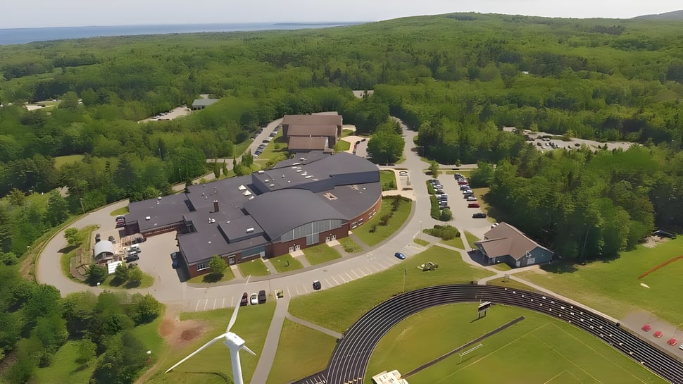 El edificio de varios pisos de la Camden Hills Regional High School se encuentra en un paisaje boscoso con una carretera de acceso y turbinas eólicas al fondo.