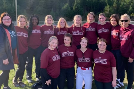 Un grupo de alumnas de la Carbonear Collegiate High School está de pie en un campo de hierba rodeado de árboles.