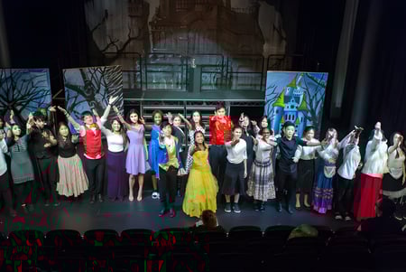 Un grupo de estudiantes del Cardiff Sixth Form College presenta una actuación en el escenario con coloridos disfraces frente a un fondo de bosque.