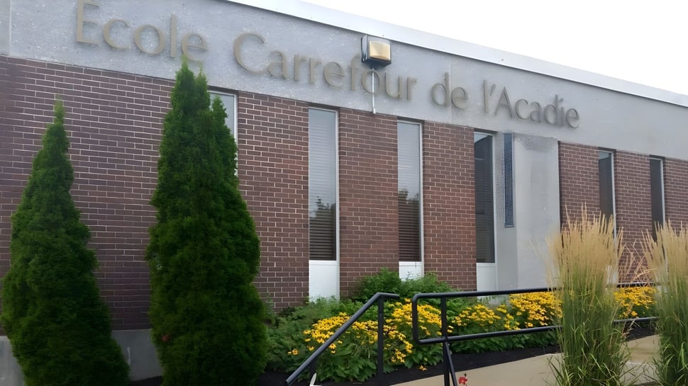 El edificio de ladrillo de la escuela Carrefour de l’Acadie está rodeado de altos árboles perennes y plantas en flor.