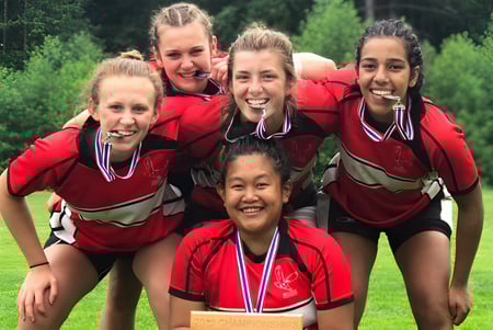 Un grupo de jóvenes mujeres en uniformes rojos con medallas está de pie en un campo en el terreno de la Carson Graham Secondary School.