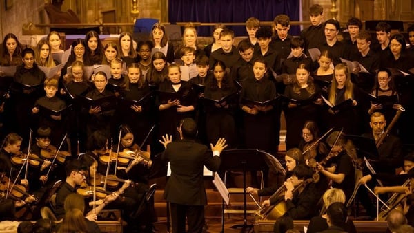 Un gran coro en túnicas negras canta en el escenario con una orquesta al frente en la Caterham School.