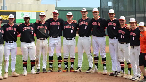 Estudiantes de la Cathedral Preparatory School están en uniforme de béisbol con cascos en el campo de béisbol frente a un edificio.