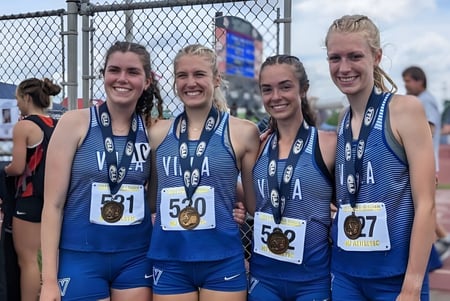 Cuatro estudiantes de la Cathedral Preparatory School están en uniformes deportivos junto al estadio de atletismo cercado.