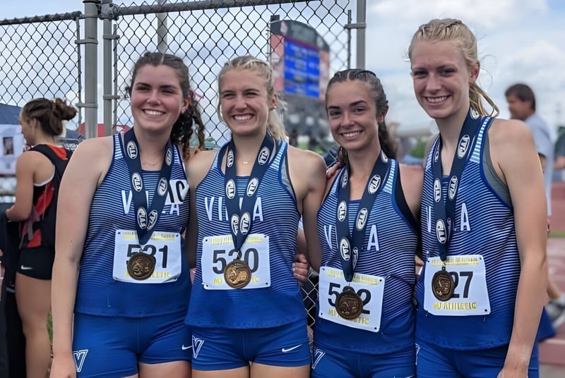 Cuatro estudiantes de la Cathedral Preparatory School están en uniformes deportivos junto al estadio de atletismo cercado.