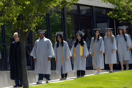 Las graduadas y graduados de la CATS Academy Boston están de pie en túnicas blancas en un campo frente a un edificio.