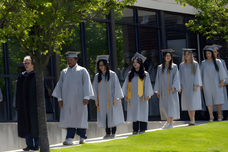 Las graduadas y graduados de la CATS Academy Boston están de pie en túnicas blancas en un campo frente a un edificio.