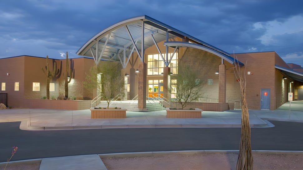 El moderno edificio con fachada de vidrio y techo curvado en el terreno del Cave Creek Unified School District está rodeado de árboles.
